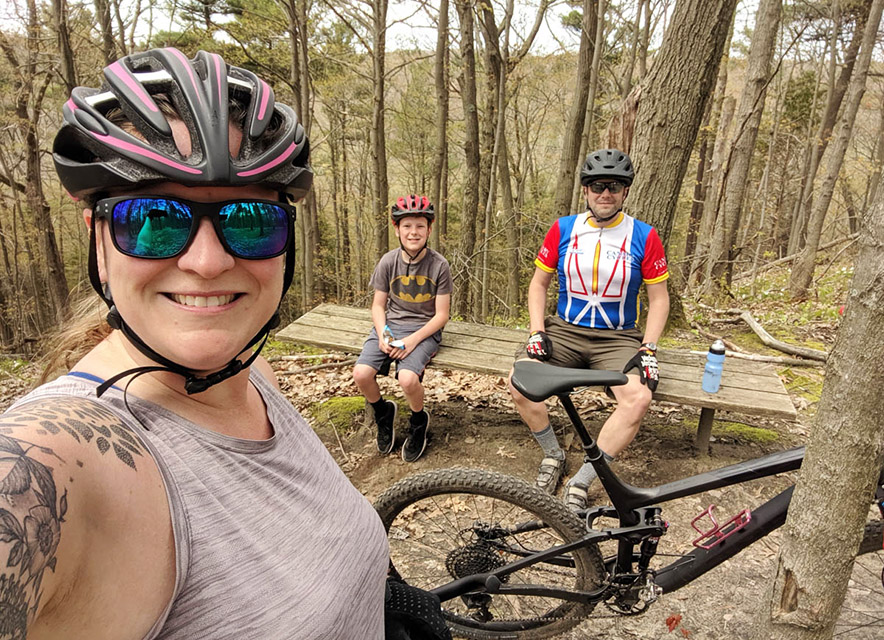 Sues Outdoor Crew Woman with bicycle helmet on taking selfie with husband and son in background sitting on picnic table in the woods